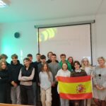 uczniowie wraz z prowadzącą wykład pozujący do zdjęcia, jedna osoba trzymająca flagę Hiszpanii