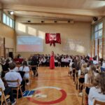 uczniowie siedzący na sali gimnastycznej podczas uroczystej akademii z okazji 11-go Listopada, na środku uczennica śpiewająca