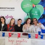 uczennice wraz z dyrektorem siedzące przy stole na tle baneru z ofertą szkoły
