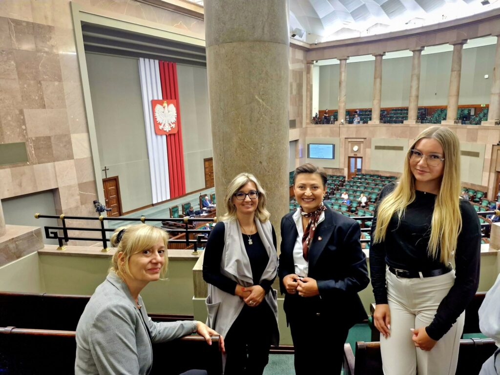 kadra pedagogiczna, poseł i uczennica stojące na tle sali posiedzeń Sejmu RP